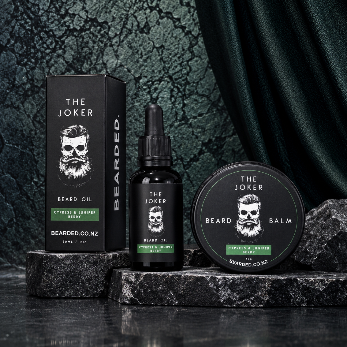 Beard grooming set by The Joker, including a beard oil bottle, a beard balm tin, and a product box, arranged on dark stone blocks against a dark textured backdrop with draped fabric.
