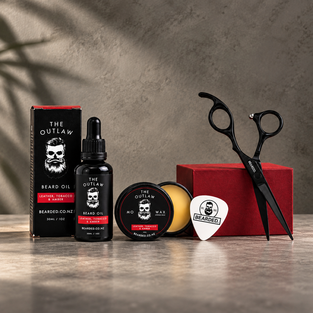Beard grooming kit The Outlaw by BEARDED. featuring beard oil, mo wax, a wax pick and black scissors, arranged on a stone surface with a textured gray background.